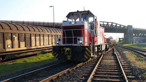 Eine rot-weiße Lokomotive, die eine Reihe von Güterwagen auf einer Eisenbahnstrecke zieht, mit einer Brücke und einigen Gebäuden im Hintergrund.