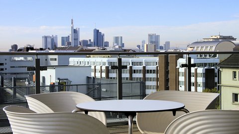 Ein moderner Balkon mit einem runden Tisch und vier weißen Stühlen mit Blick auf die Frankfurter Skyline mit hohen Wolkenkratzern.