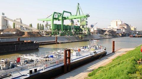 Eine Hafenszene mit einem Lastkahn mit der Aufschrift "VOLENDAM", der in der Nähe eines Krans vertäut ist, umgeben von verschiedenen industriellen Strukturen und Ausrüstungen.