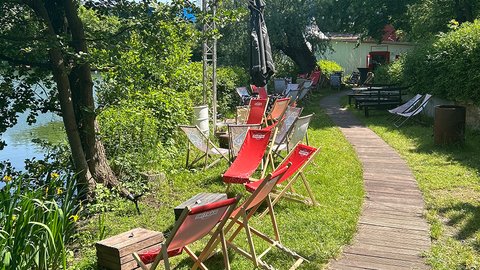 Eine ruhige Umgebung am Schedler See mit mehreren rot-weiß gestreiften Stühlen in einer Reihe, einem Holzweg und ein paar Menschen im Hintergrund.