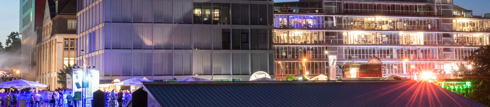 Eine lebendige städtische Umgebung am Abend, mit modernen Gebäuden, die von bunten Lichtern beleuchtet werden, und einer Menschenmenge, die sich draußen versammelt hat, um die Atmosphäre zu genießen.